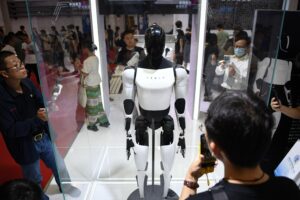 Photo of a faceless Tesla-branded humanoid robot in a glass display case surrounded by people taking pictures of the robot on their smartphones.